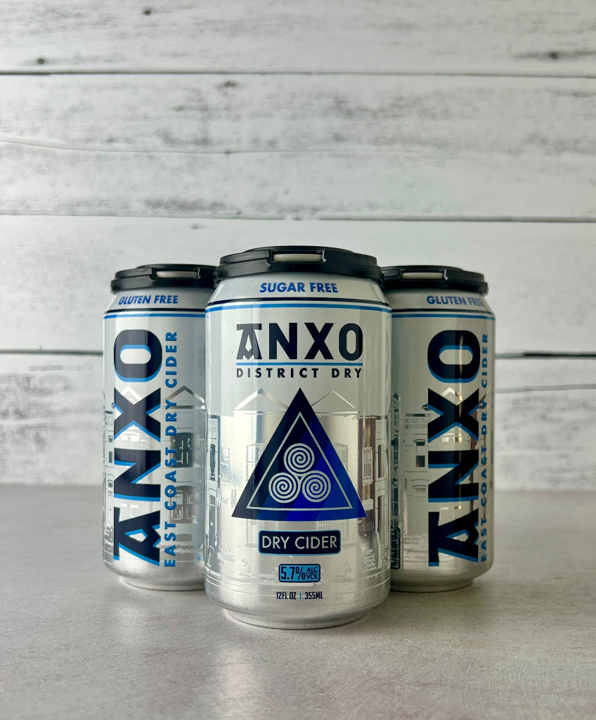 Photo of a 4-pack of  12-oz cans of Anxo District Dry cider on a gray surface with a white wooden background