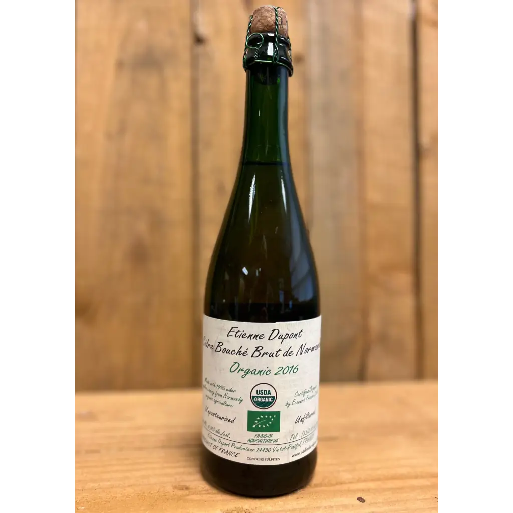 Cork and caged bottle of Etienne Dupont Cidre Bouche Brut de Normandie Organic 2016 cider on a wood table with wood background