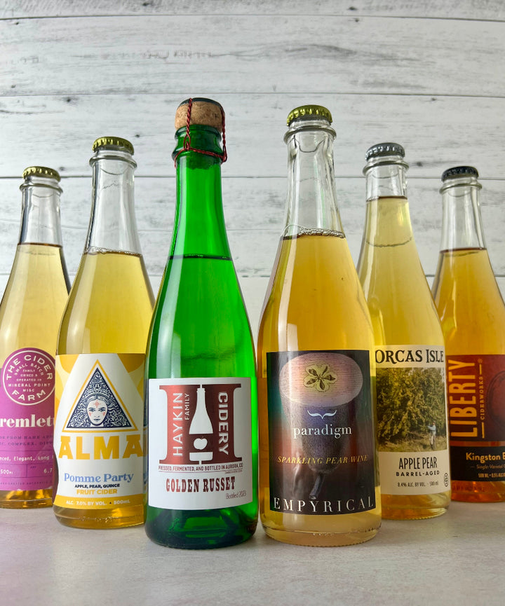 Photo of six bottles of 500 and 375 mL bottles, including The Cider Farm Tremlett's, Alma Pomme Party, Haykin Cidery Golden Russet, Empyrical Paradigm sparkling pear wine, Orcas Isle Apple Pear Barrel-Aged cider, and Liberty Ciderworks Kingston Black
