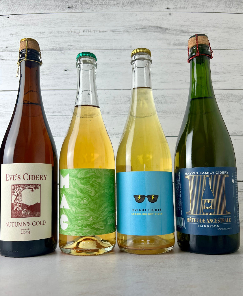 Photo of 4 750 mL bottles of cider, from left to right: Eve's Cidery Autumn's Gold, Bauman's Mac, Little Pomona Bright Lights Sparkling Dry Cider, Haykin Family Cidery Methode Ancestrale Harrison