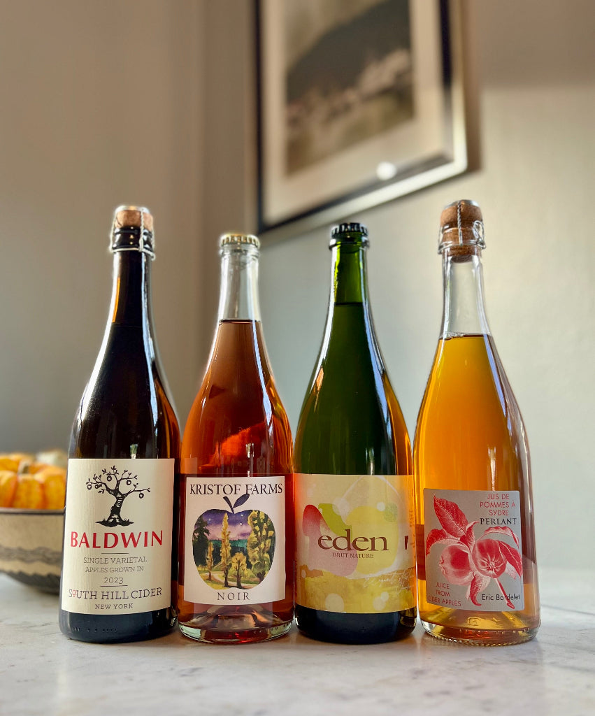 Photo of 4-bottles of cider, from left to right: South Hill Cider Baldwin, Kristof Farms Noir, Eden Brut Nature, Eric Bordelet Perlant Juice from Cider Apples