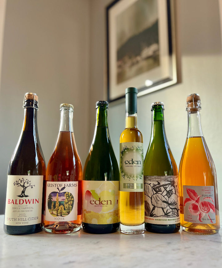Photo of 6-bottles of cider, from left to right: South Hill Cider Baldwin, Kristof Farms Noir, Eden Brut Nature, Eden Heirloom Ice Cider, PIF Giganticus Maximus Keeved Cider, Eric Bordelet Perlant Juice from Cider Apples