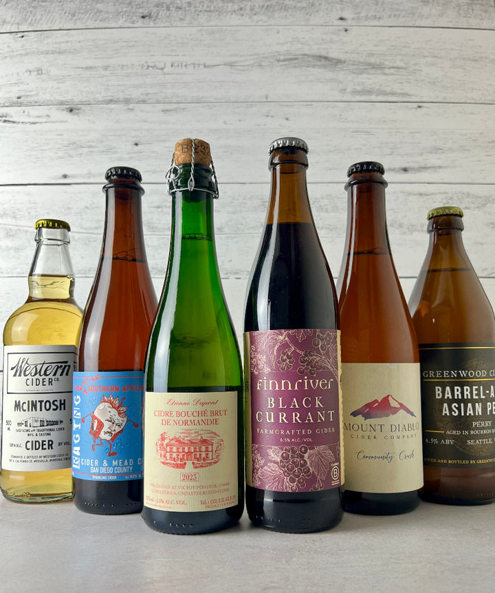 Photo of six 500 mL bottles of cider, from left to right: Western Cider McIntosh, Raging Cider & Mead Them Pet-Nat Southern Apples, Etienne Dupont Cidre Bouche Brut de Normandie, Finnriver Black Currant, Mount Diablo Community Crush, and Greenwood Cider Barrel-Aged Asian Pear