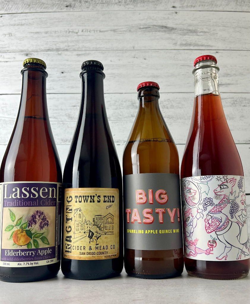 Photo of 4-bottles of cider, from left to right: Lassen Elderberry Apple, Raging Cider Town's End, Durham Big Tasty!, and Two Broads Cider I Feel Pretty