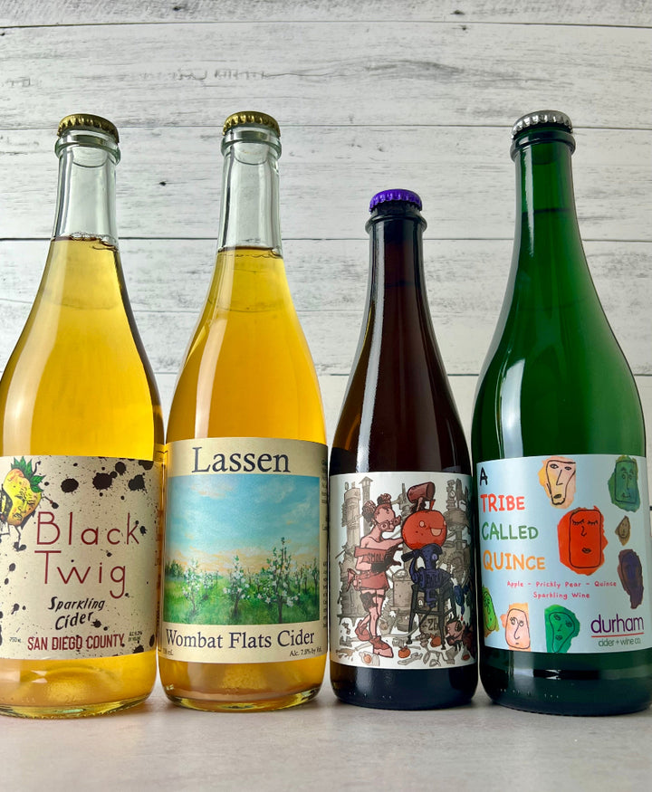 Photo of 4-bottles of cider, from left to right: Raging Cider Black Twig, Lassen Cider Wombat Flats, Two Broads Cider Wild Heirloom, and Durham Cider A Tribe Called Quince