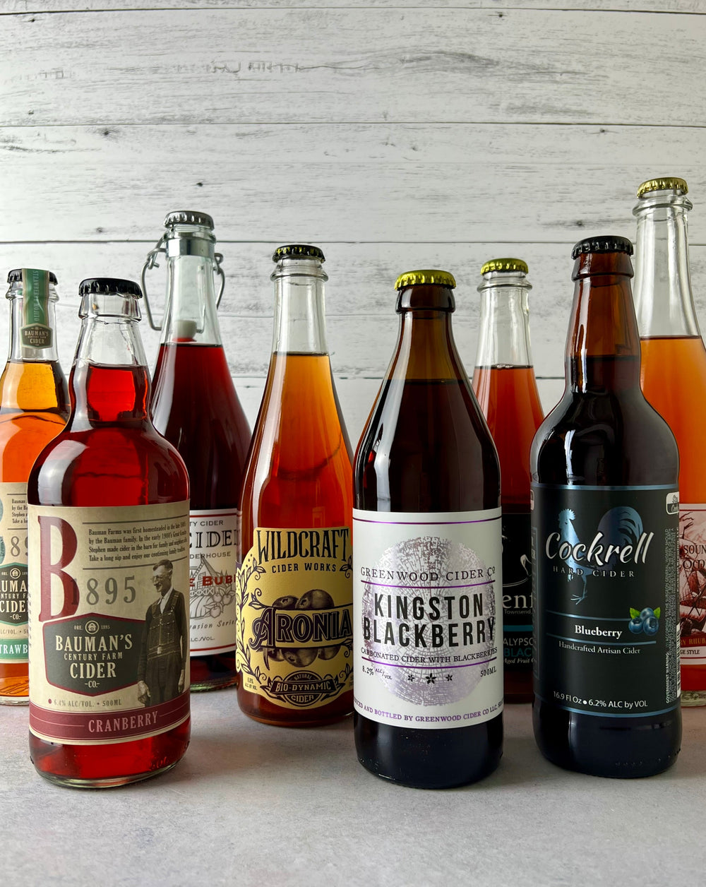 Multiple bottles of fruited cider, including Bauman's Strawberry Mojito, Bauman's Cranberry, Sea Cider Bramble Bubbly, Wildcraft Aronia, Greenwood Kingston Blackberry, and Cockrell Blueberry