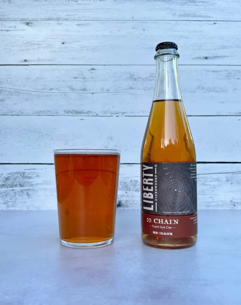 500 mL bottle of Liberty Ciderworks 55 Chain - English Style Cider next to a glass full of amber-colored cider