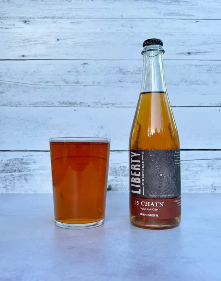 500 mL bottle of Liberty Ciderworks 55 Chain - English Style Cider next to a glass full of amber-colored cider