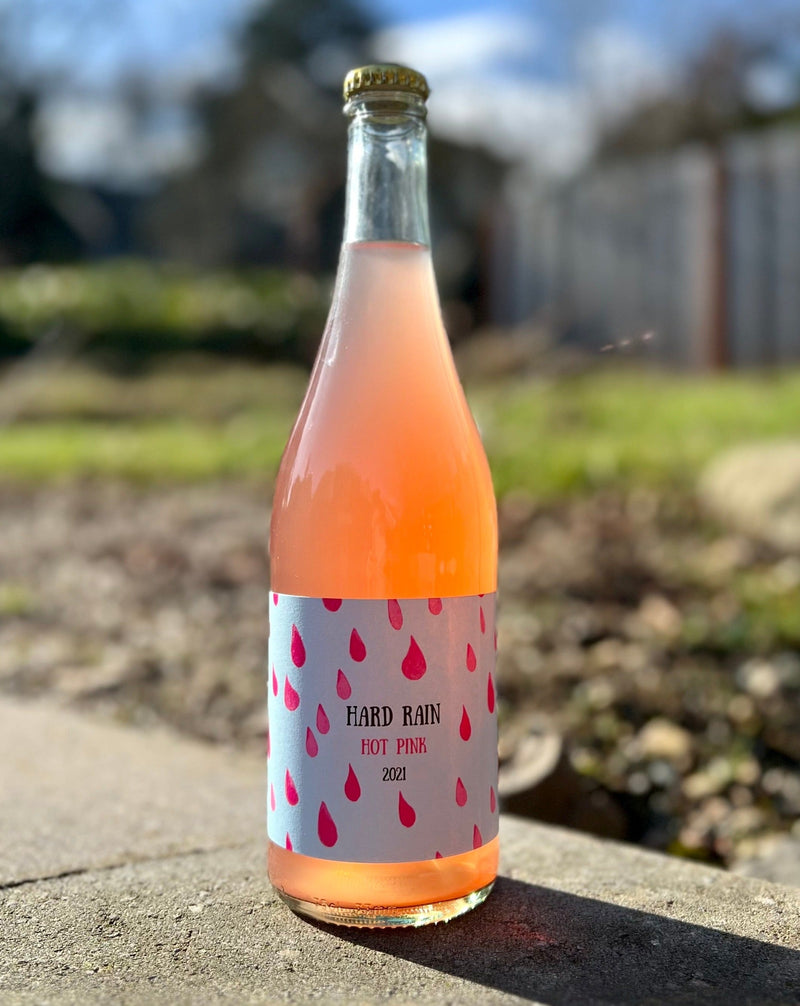 Bottle of Little Pomona Hard Rain Hot Pink 2021 Cider in a clear bottle, showing off the cloudy pink color of the cider