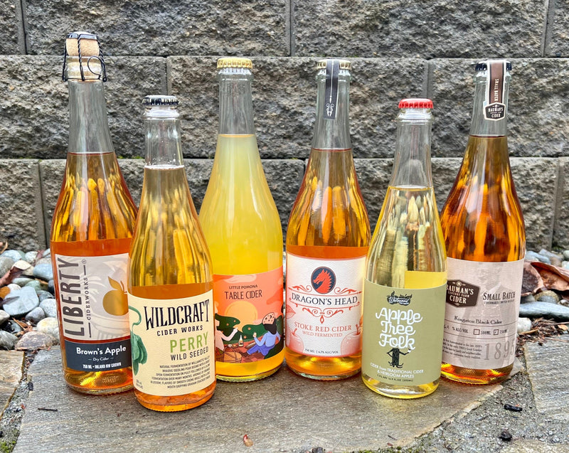 6 Bottles of cider, from left to right: Liberty Brown's Apple, Wildcraft Perry, Dragon's Head Stoke Red Cider, Whitewood Apple Tree Folk, and Bauman's Kingston Black cider